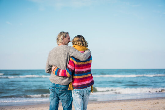 ture couple standing on the beach with arms around, looking at the sea