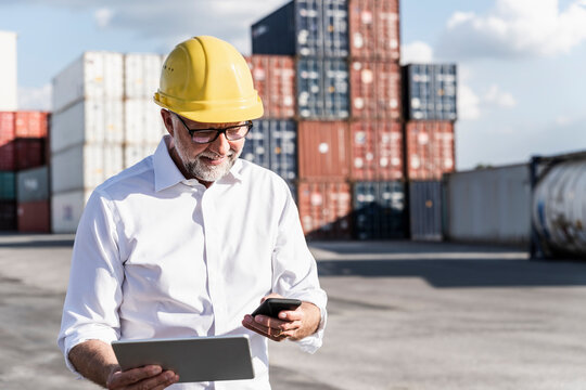 Businessman at cargo harbour, wearing safety helmet, using smartphone and digital tablet