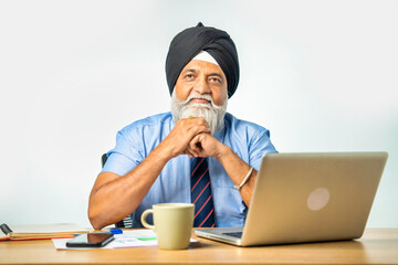 Confident senior Indian Sikh man at table with laptop, resting hands, success gesture or pointing