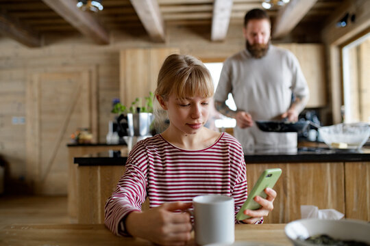 Girl is sitting at the table, drinking tea and scrolling on her smartphone. Morning routine for family.