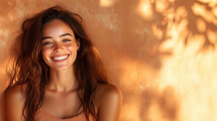 Fototapeta premium A smiling woman with long hair and freckles enjoys sunlight, leaning against a warm-toned wall, with shadows of leaves playing across her face.
