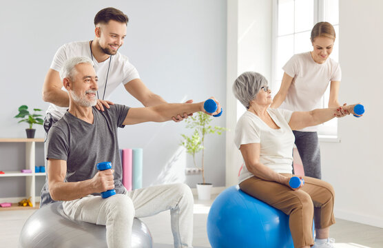 Happy senior people doing physiotherapy exercises with sports equipment. Smiling physiotherapists helping old man and woman who are sitting on fitness balls and holding dumbbells