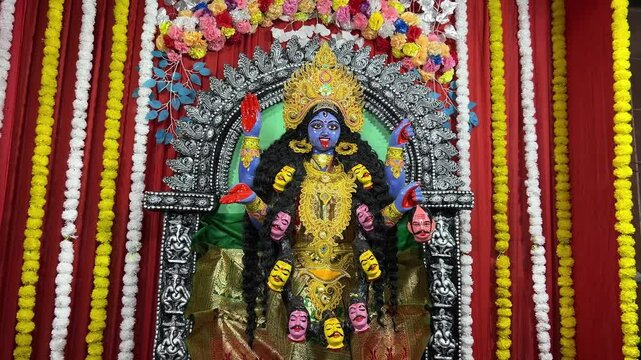 A dynamic shot of the Maa Kali idol adorned in blue, with a beautiful arrangement of red, yellow, and white strings, set against a vibrant red backdrop.