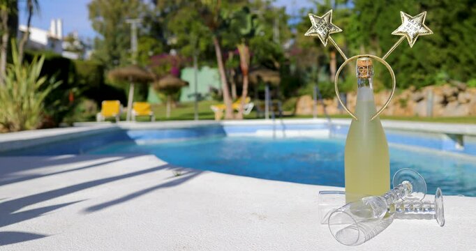Footage of a champagne bottle with a head band bopper saying happy New Year surrounded by two wine glasses on a sunny day by a swimming pool indicating New Years Eve in the sun