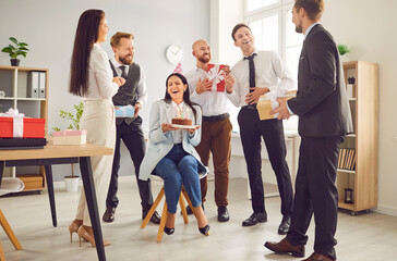 Group of young smiling business people congratulating their happy female colleague on her birthday giving her birthday cake and present gift boxes on workplace in office. Corporate party