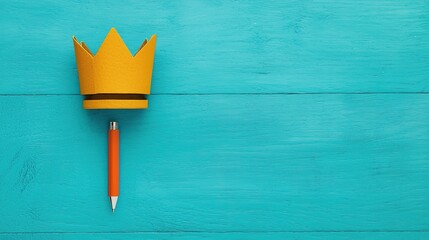 A bright yellow crown sits atop an orange pen against a teal wooden background, creating a playful and creative atmosphere.