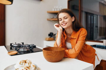 A cheerful young woman in an orange shirt enjoying cooking in a modern kitchen, smiling at a wooden bowl, with a warm and inviting atmosphere