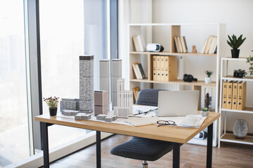 Modern office desk features architectural model and laptop setup. Bright workspace includes plants, files, and glasses. Ideal for conceptualizing urban design projects with contemporary elements.