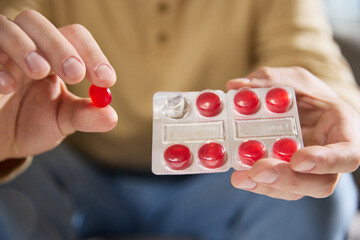 Close up photo of man in casual beige shirt holds blister pack of red lozenges, sitting in bright,...