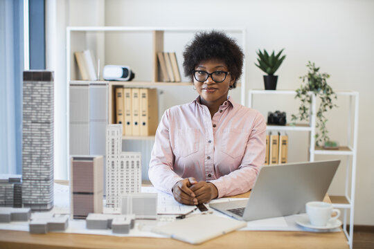Young African woman architect designing modern city model at desk. Professional workplace with laptop, blueprints, and coffee. Creative architectural process focuses on urban development. - Powered by Adobe