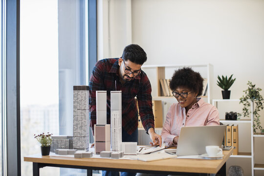 Indian man and African woman collaborate on architecture project with city models. Office setting with teamwork and creativity enhancing urban planning. Professional environment showcasing design.