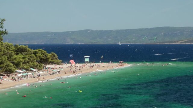 Crowds of tourists sunbathe, swim, and relax on Zlatni Rat Beach on the island of Brac (Brač), Croatia. A vacation on the Adriatic Sea coast during the tourist season. Waves crash against the shore.