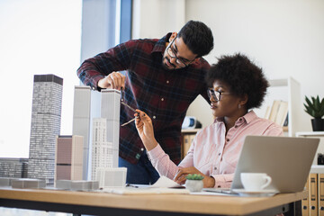 Indian man and African woman architects collaborate on model project for modern city. Diverse teamwork in bright office setting with creative discussion and urban planning focus.