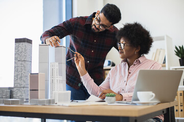 Indian man and African woman architects collaborate on model project for modern city. Diverse teamwork in bright office setting with creative discussion and urban planning focus.