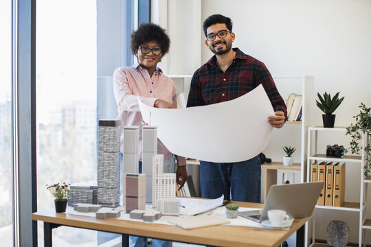 Indian man and African woman architects analyzing blueprints for city development project. Creative teamwork in modern office setting. Collaborative work with model buildings, plants, and city view. - Powered by Adobe