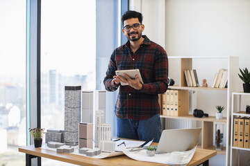 Fototapeta premium Indian male architect develops modern city project using digital tablet in creative workspace. Tall building models on desk symbolize innovative urban design. Age group young adult to middle-aged.