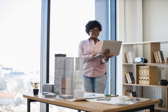 African American female architect in casual attire develops building project using laptop. Bright workspace with architectural models and natural light enhances creative process.