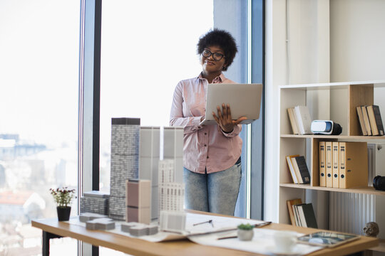 African American female architect in casual attire develops building project using laptop. Bright workspace with architectural models and natural light enhances creative process.