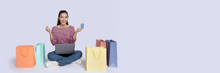 A happy shopper sits on the floor surrounded by colorful shopping bags while holding a credit card...