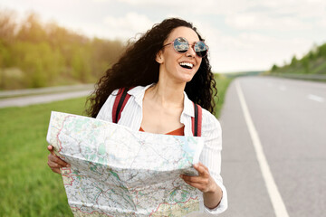 Joyful millennial lady walking along asphalt road with map, choosing travel destination,...