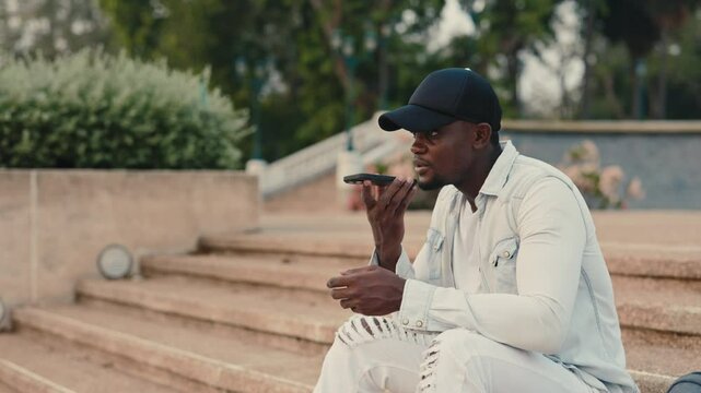 Portrait of African American man recording voice message on smartphone in city park. Young guy uses mobile phone sitting on stone stairs. Modern busy urban lifestyle