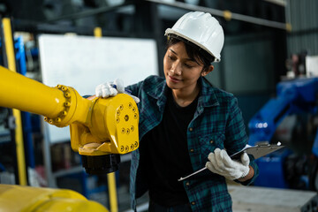 Female engineer wearing a safety helmet and gloves carefully examines a yellow robotic arm while...