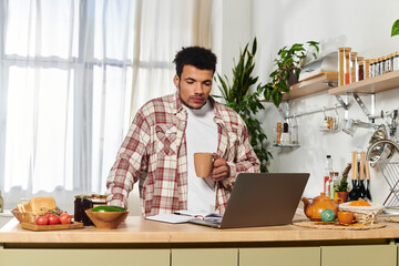 A young handsome man is sipping coffee while focused on his laptop in a bright kitchen.