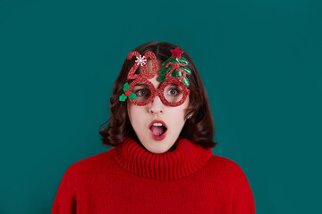 A surprised girl in colorful festive glasses in the shape of a Christmas tree and the number 2025. A bright red sweater creates a festive atmosphere. 