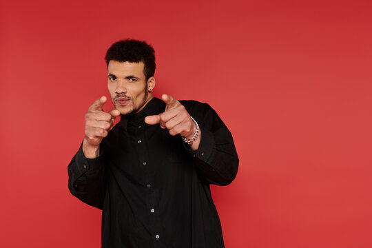 A young and handsome man strikes a playful pose while smiling against a bold red wall.