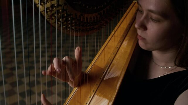 Young Caucasian female harpist plucking masterfully strings on wooden harp in dim lit hall with checkerboard floor