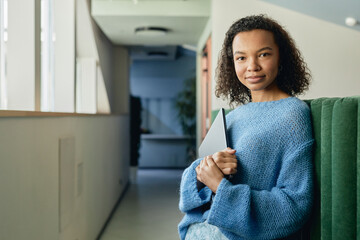 Portrait of young multiethnic woman wearing blue sweater posing for camera with laptop in hands...