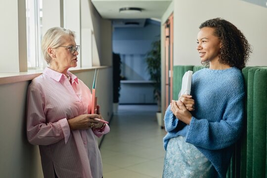 Side view of mature businesswoman casually chatting with younger employee of multiethnic background establishing intergenerational contact in workplace while standing in hallway, copy space