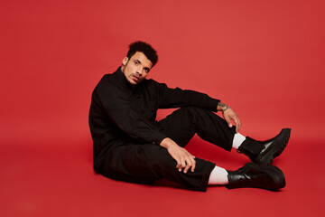 A stylish young man sits on the ground, projecting confidence with a striking red background.