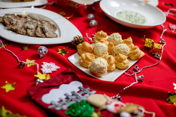 A plate of golden cream puffs with creamy filling, arranged on a festive table decorated with Christmas ornaments and a red tablecloth.