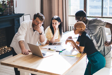 Creative multiracial people seriously looking at laptop at table with drawings