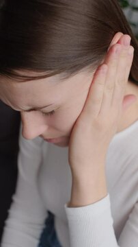 Vertical video of sad young brunette female holding painful ear, suddenly feeling strong ache. Unhealthy caucasian woman suffering from painful otitis sitting on couch at home. Health problems concept