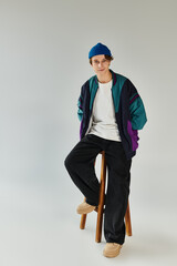 A handsome young man sits on a stool, showcasing his stylish bomber jacket in a studio.