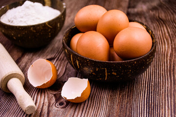 chicken eggs in a deep wooden bowl on the background of a wooden table.  two shells of broken eggs in the foreground, flour in a bowl and a wooden rolling pin. Composition for creating a homely atmosp