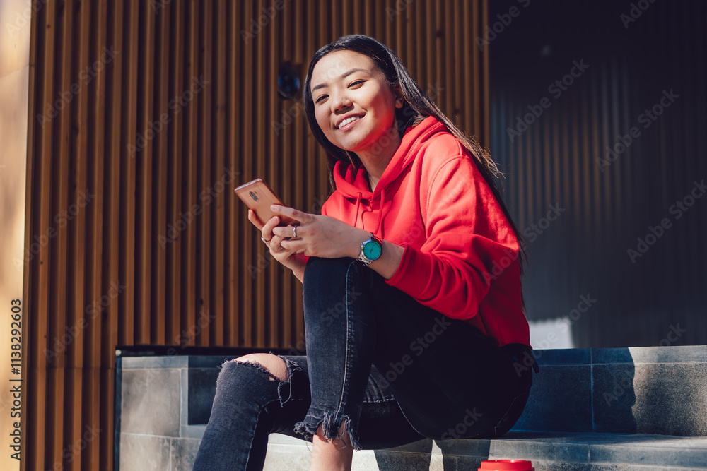 Poster portrait of cheerful chinese hipster girl with cellular device in hand resting at urban stairs durin
