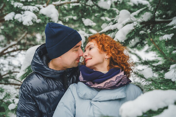 Fototapeta premium Love romantic love story. Happy couple of middle aged millennials, bright red-haired woman in winter snow park. Romantic date, kissing, hugging. Walking, having fun. Stylish blue clothes, jacket, hat