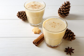 Tasty Christmas cocktail (eggnog) in glasses, pine cones and spices on white wooden table, closeup. Space for text