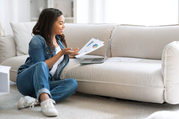 A young woman is sitting on the floor near a sofa, reviewing documents while using her laptop. She...