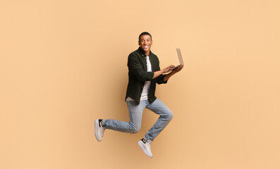 A smiling African American man in casual attire jumps in excitement while holding a laptop in front of a warm, solid backdrop. His energy reflects a positive lifestyle and motivation.