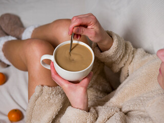 Woman in cozy bathrobe relaxes on bed, savoring warm cup of coffee and stirring it slowly. Fresh tangerines nearby enhance tranquil atmosphere as she enjoys peaceful morning with her cup of coffee