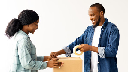 Fototapeta premium Moving Day. Cheerful African American Spouses Packing Belongings In Cardboard Box While Relocating To Their New Apartment, Happy Young Black Couple Using Scotch Tape To Make Package, Closeup Shot