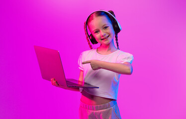 A preteen girl with braided hair smiles while wearing headphones and pointing at her laptop. Neon...