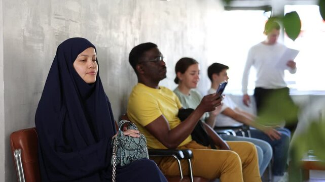 Girl in traditional veil, along with other people of different nationalities, is waiting for a specialist sitting on a chair in the hall