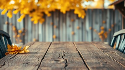 A weathered wooden table with a single yellow leaf resting on its surface, set against a backdrop of a blurred autumnal scene with a fence and a glimpse of foliage.