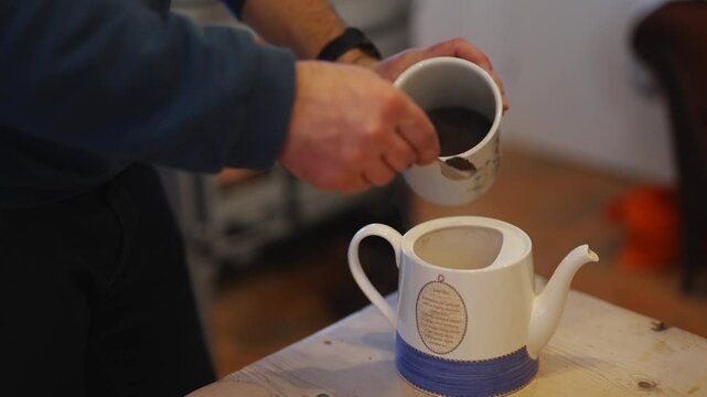 A man is pouring coffee into a white cup. The scene is set in a cozy living room with a fireplace in the background