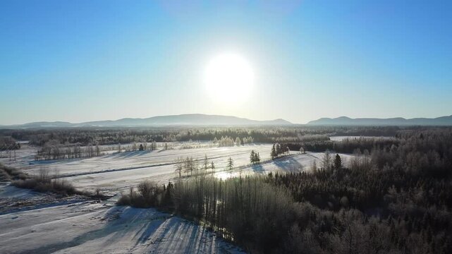 Winter aerial panorama of a snow-covered rural landscape under bright sunlight and a vivid blue sky with the Appalachian Mountains in the distance. Sayabec, Matapedia Valley, Quebec, Canada.